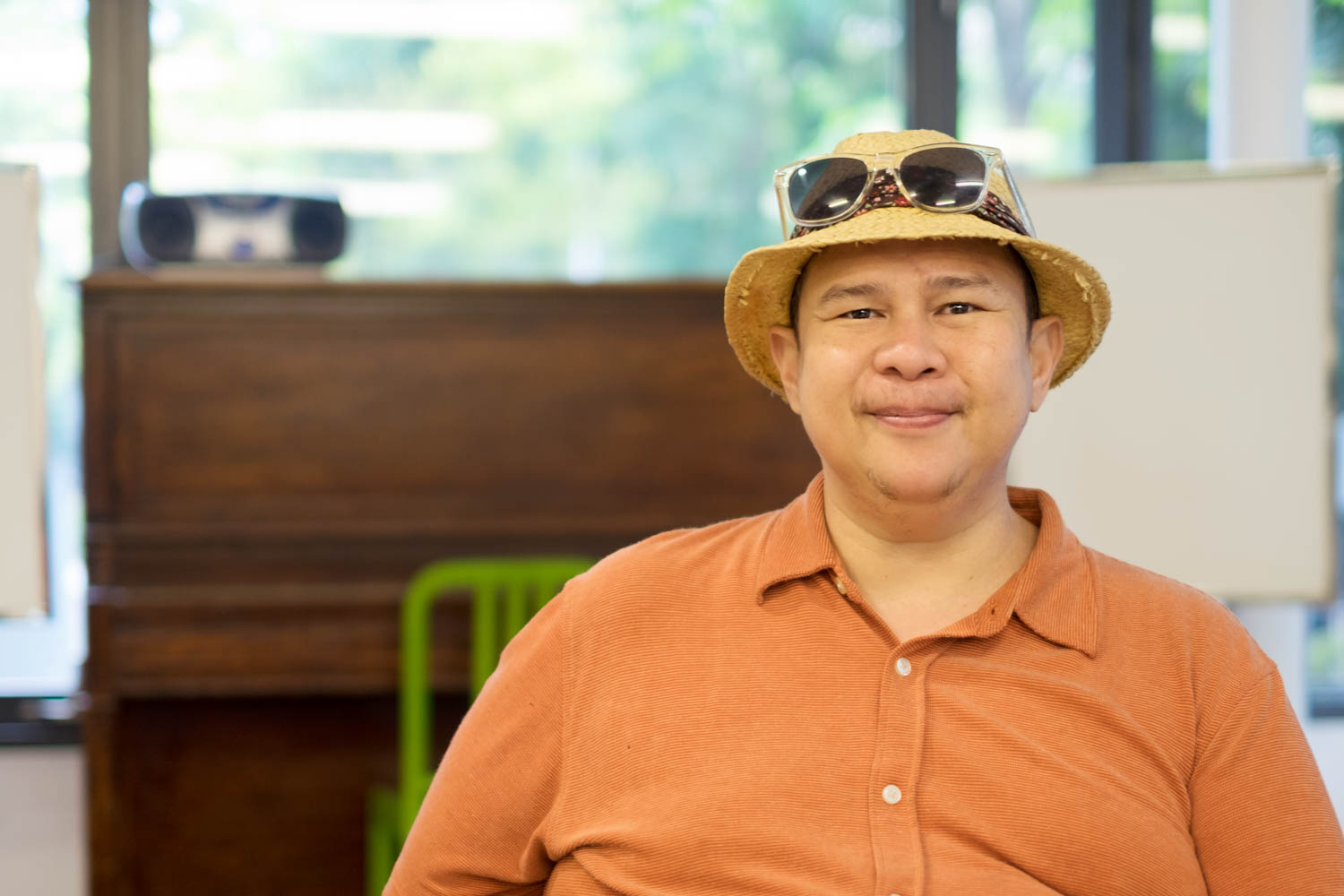 Lawrence sitting in a chair with a table in front, wearing an orange buttoned and collared shirt, a straw hat and black sunglasses propped up on the hat. Lawrence is looking at the camera and smiling. There is a blurred radio in the background.