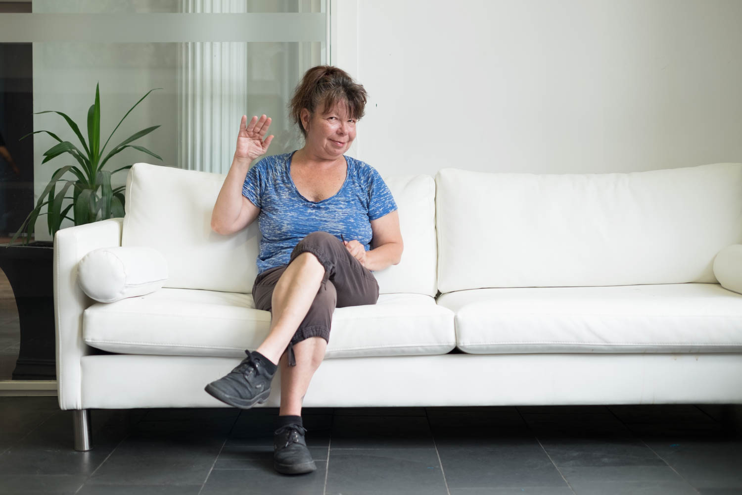 Colleen wearing a blue T-shirt, brown capris, and black laced shoes sitting on a white couch and waving to the camera. Colleen has their hair in a pony tail  and there is a white pillar and a potted plant in the background.