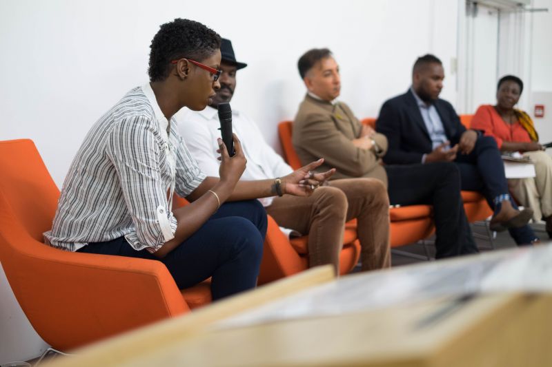 Side mid-shot of Latoya speaking in a panel on an orange sofa with a mike in hand. Four other panelists in the background sitting and listening. 