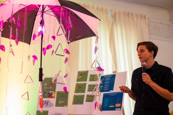 A person holding a mic and talking, in front of them an artistic installation of an umbrella with different words and cristal pipes hanging.