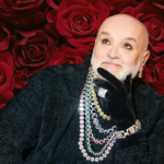 Salah Bachir wearing a Black velvet outfit and lots of dazzling diamond chains. He has his hand to his chin and is looking up and smiling